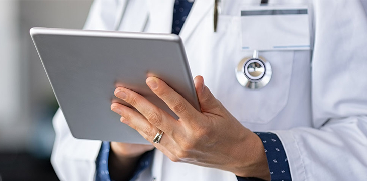 Closeup of female doctor in lab coat and stethoscope holding digital tablet, reading patient report.